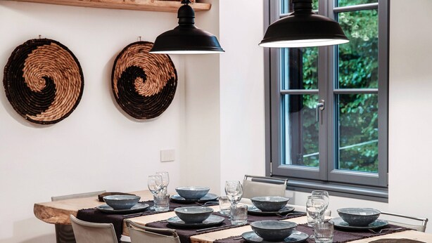 Een eetgedeelte met een houten tafel voor zes personen, zwarte borden, glazen en geweven wanddecoraties.