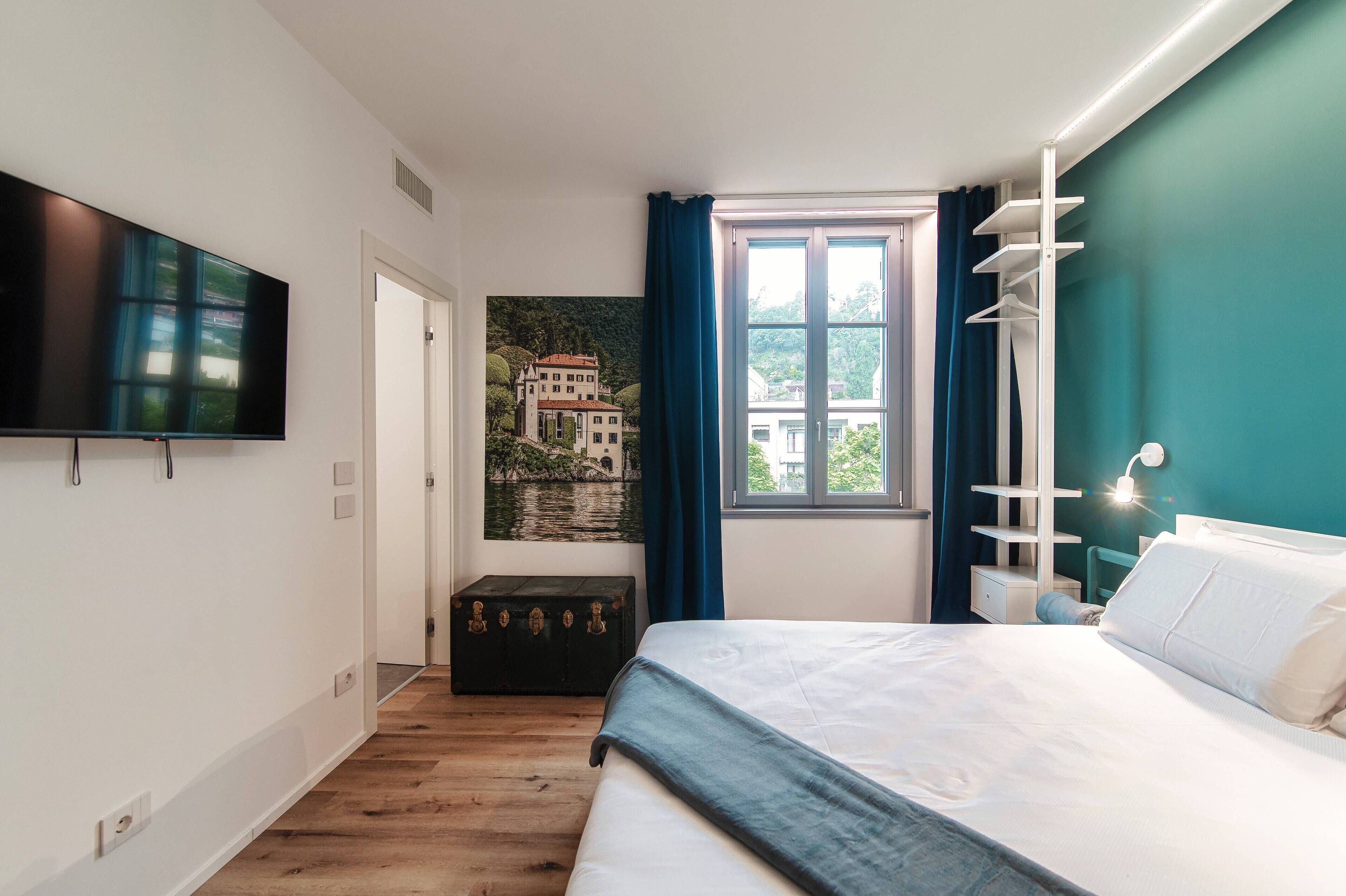 Ein Schlafzimmer mit einem Bett, einem an der Wand montierten Fernseher, einem Fenster mit Vorhängen, einem Gem älde und einer kleinen Kommode.