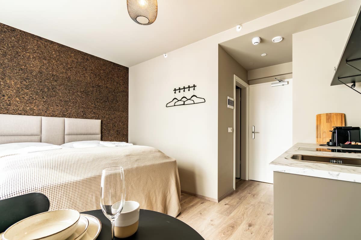 A modern hotel room with a bed, a dining table with plates and a glass, a kitchen area with a sink, and a wall-mounted coat rack.