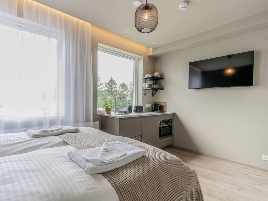 A modern hotel room with a large bed, a flat-screen TV, a kitchenette, and a window with sheer curtains.