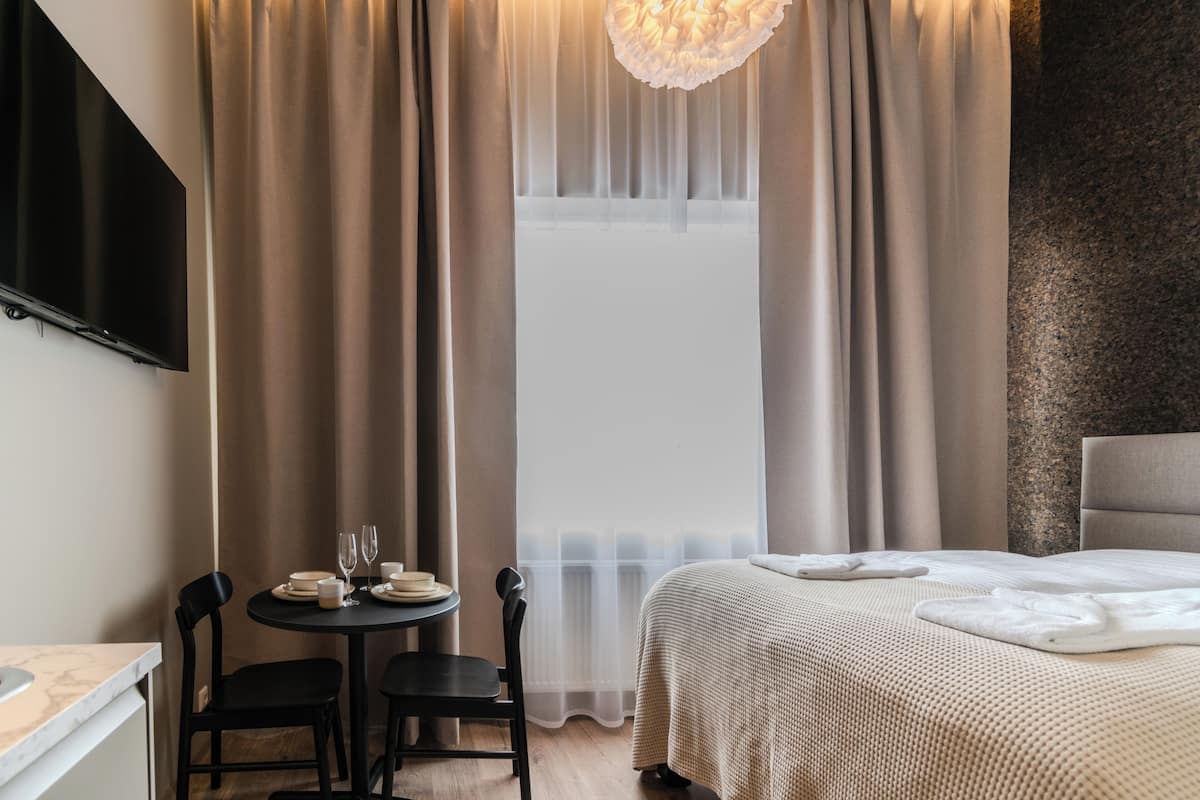 A hotel room with a bed, a small table with chairs, a TV mounted on the wall, and a window with curtains.