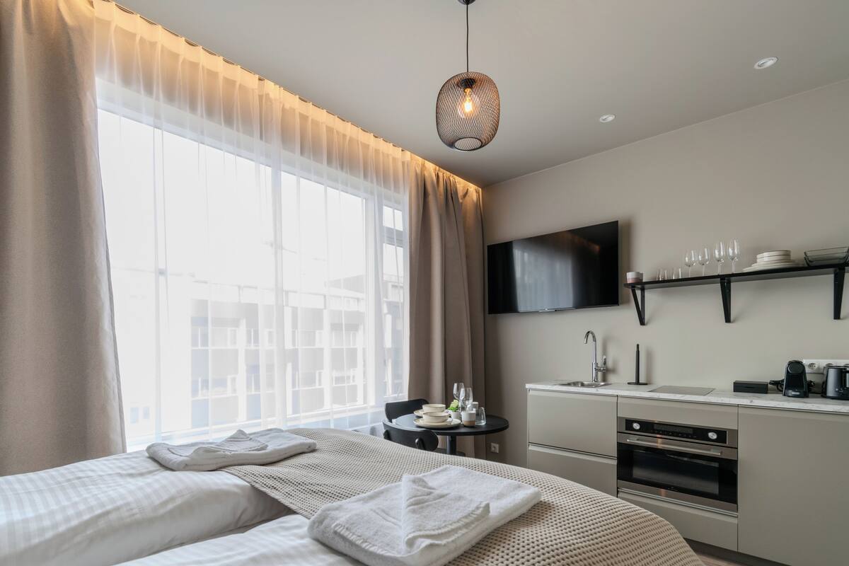 A hotel room with a bed, a small table with a TV, a kitchenette, and a large window with curtains.
