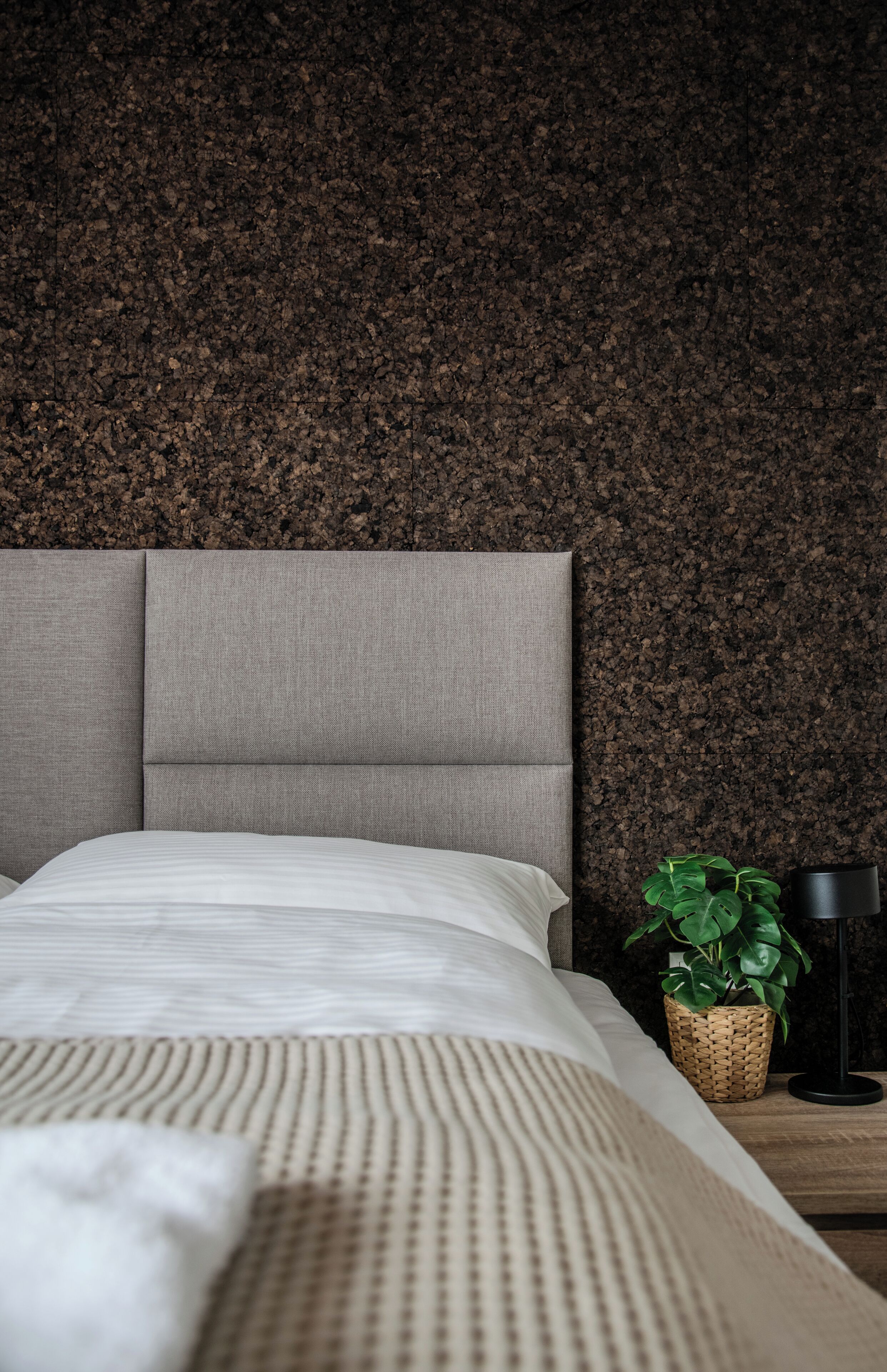 A bedroom with a textured wall, a bed with a grey headboard, a white comforter, a woven basket with a plant, and a black floor lamp.
