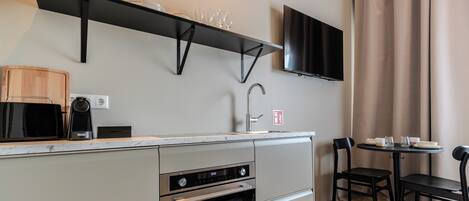 Een kleine keuken met een inbouwoven, een magnetron en een koffiezetapparaat. Er is een eethoek met een tafel en stoelen, en een flatscreen televisie aan de muur bevestigd.