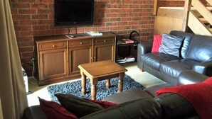 A living room with a brick wall, a television mounted above a wooden cabinet, a leather sofa, and a wooden coffee table.
