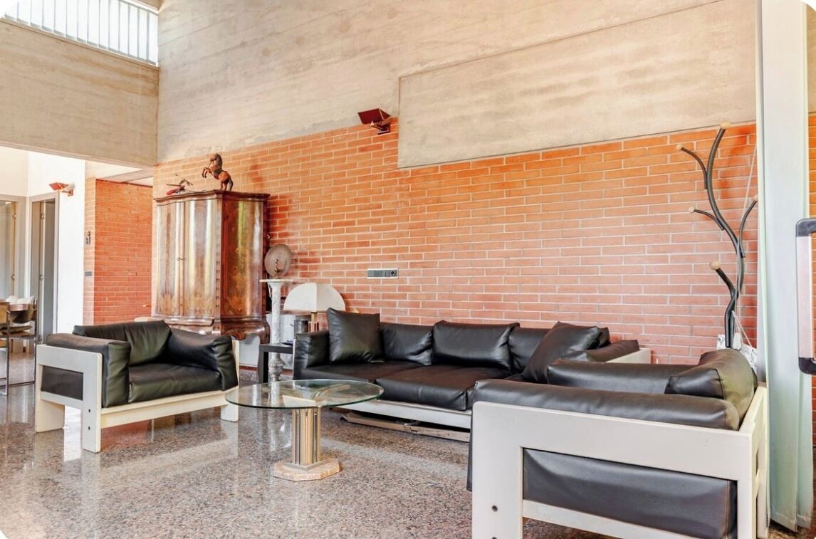 A spacious living area with a brick wall, a black leather sofa, a glass coffee table, and a white armchair.