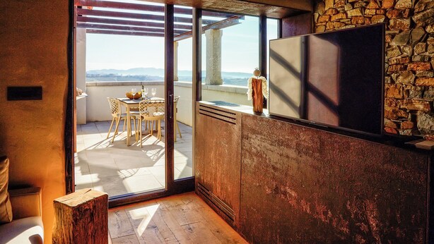 Una habitación con pared de piedra, suelo de madera, puerta de cristal corredera que da a un balcón con mesa y sillas, y un televisor de pantalla plana montado en la pared.