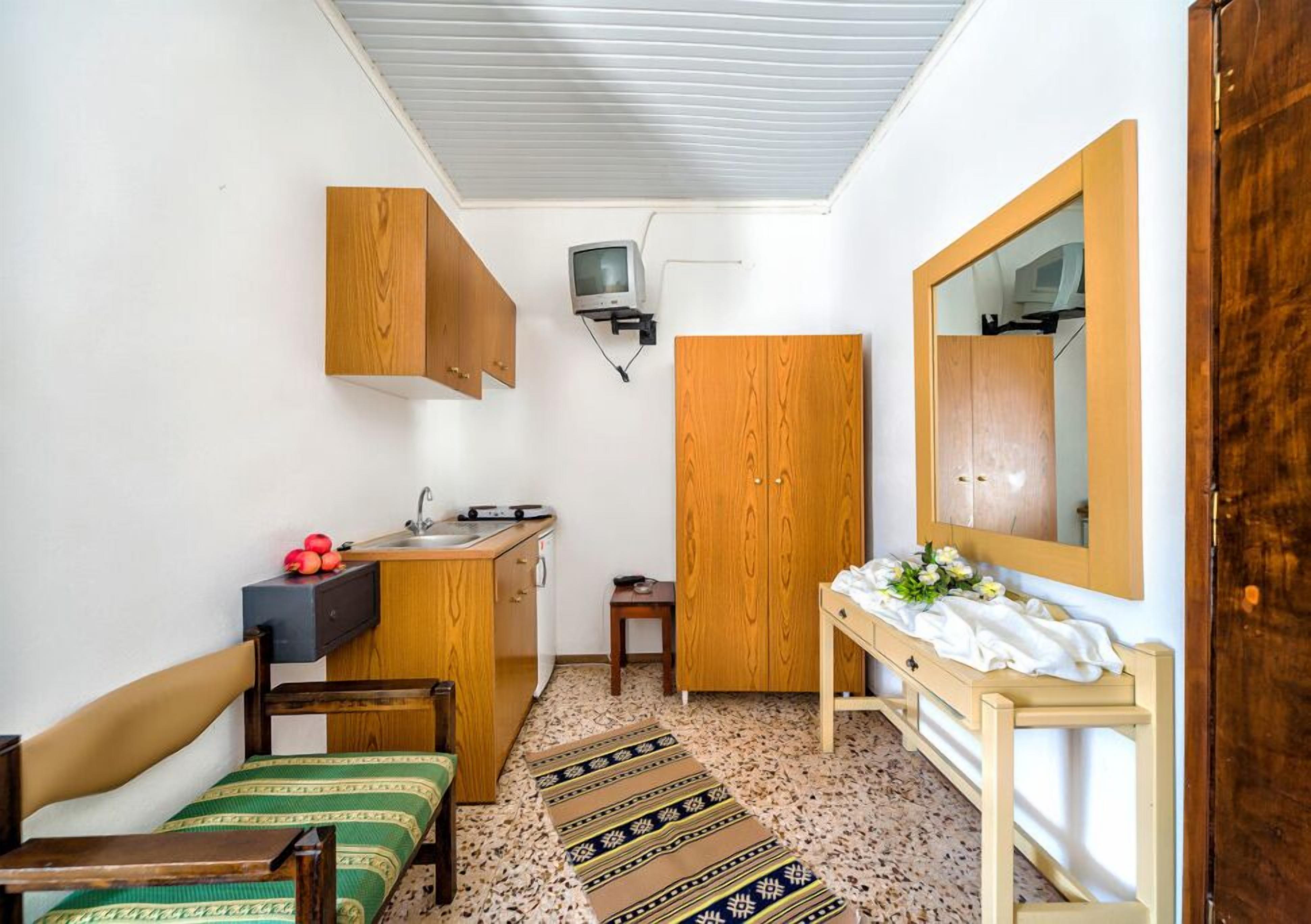 A compact room with a bed, a small desk, a mirror, a television, and a kitchenette.