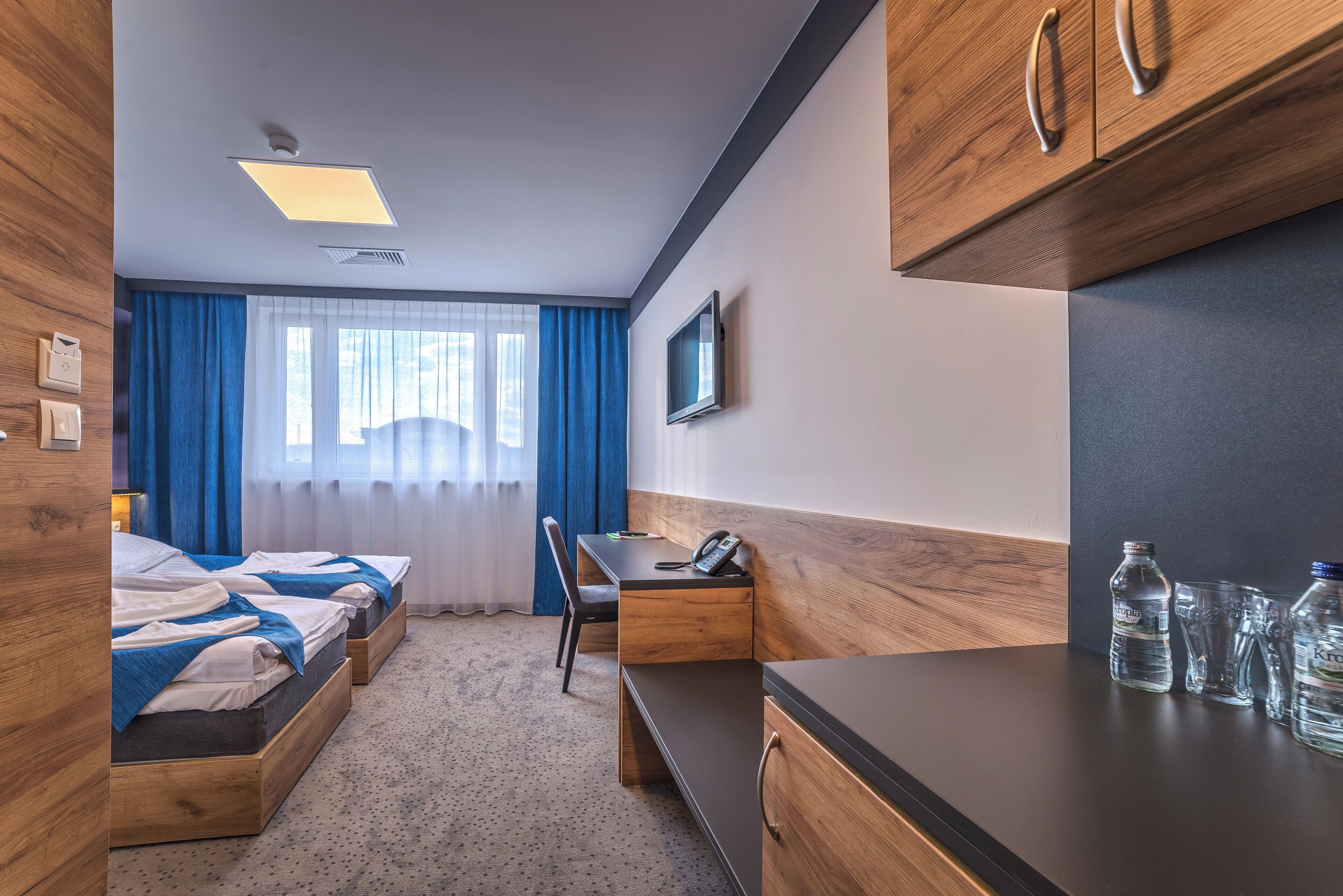 A hotel room with two beds, a desk, a chair, a TV, and a kitchenette area.