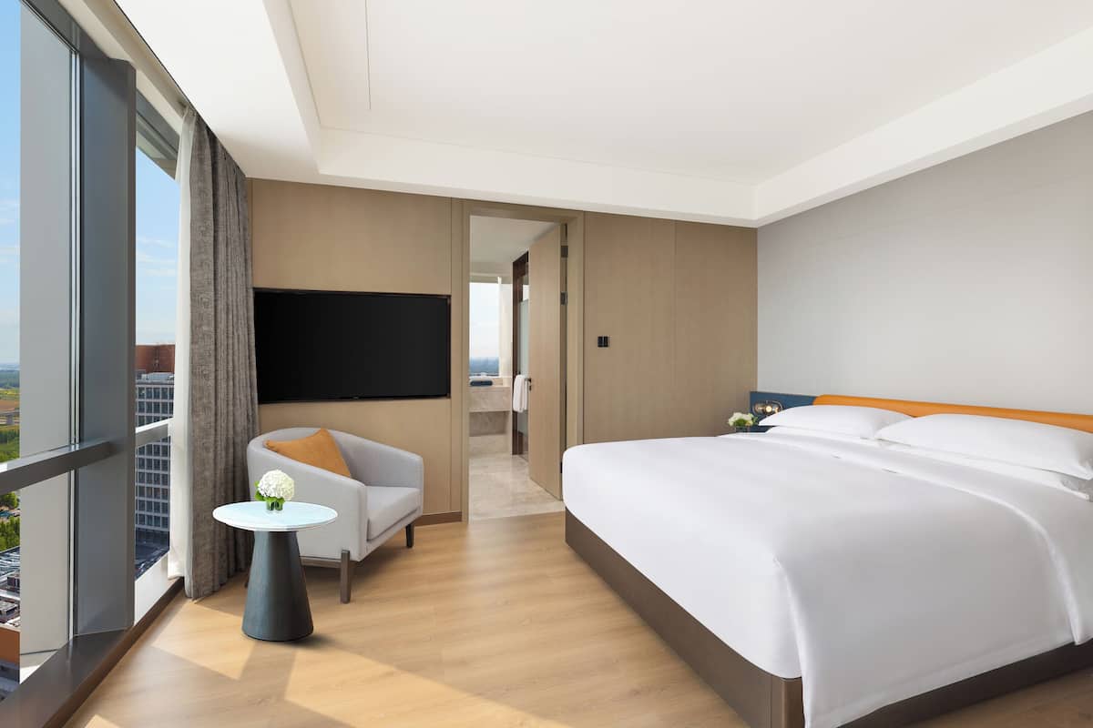 Een moderne hotelkamer met een groot bed, een zithoek met een klein tafeltje, een flatscreen televisie en uitzicht op de stad.