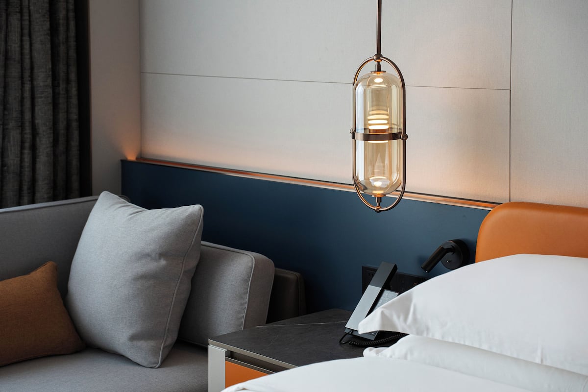 Een moderne hotelkamer met een grijze bank, een nachtkastje met een telefoon en een hanglamp.