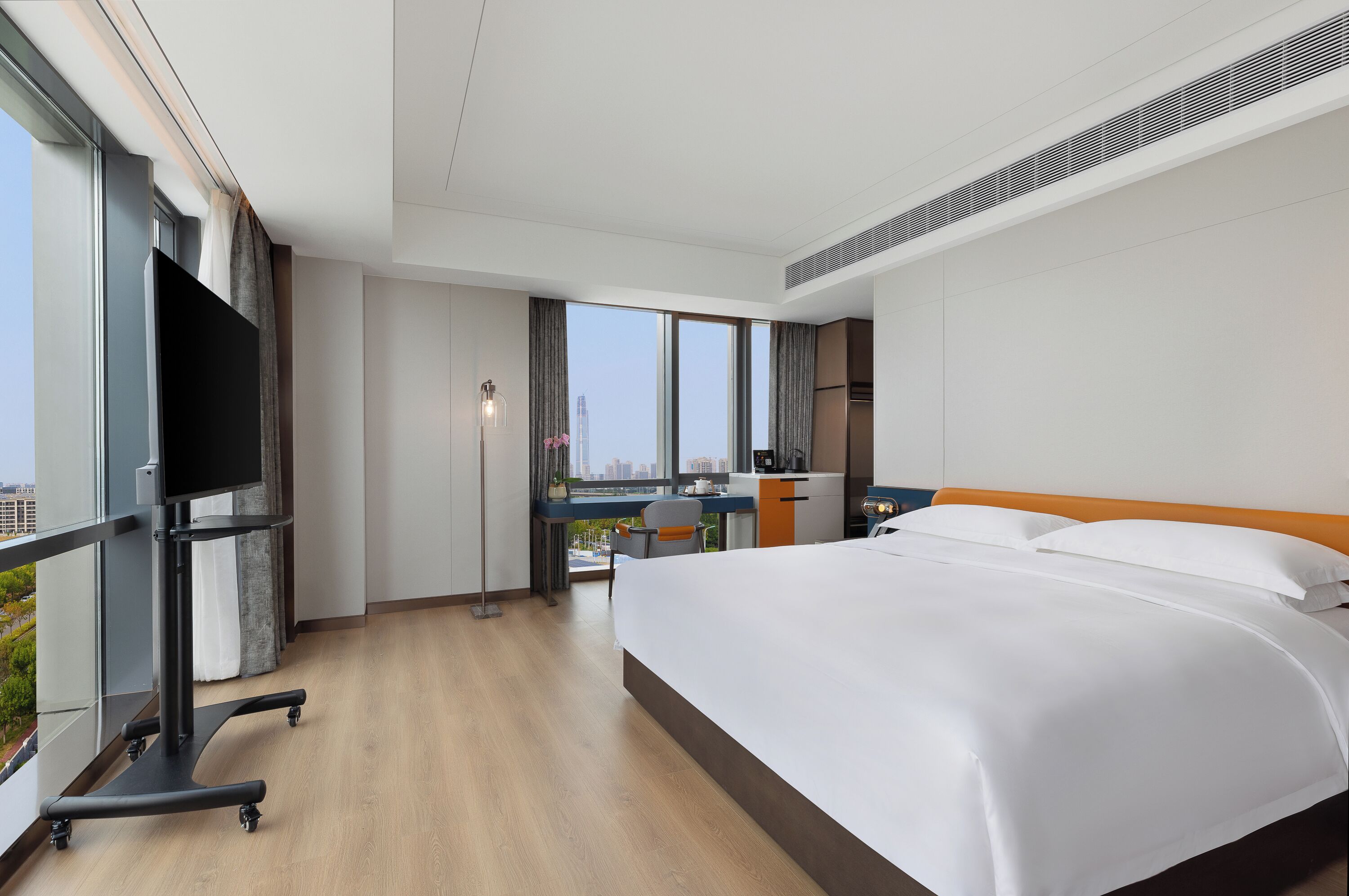 A modern hotel room with a large bed, a desk with a chair, a TV on a stand, and a city view through the windows.