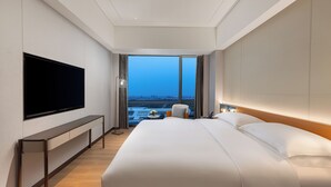 A modern hotel room with a large bed, a flat-screen TV mounted on the wall, a small table with a lamp, and a view of the cityscape through large windows. - The Perennial Tianjin, A JdV by Hyatt Hotel (Tianjin)