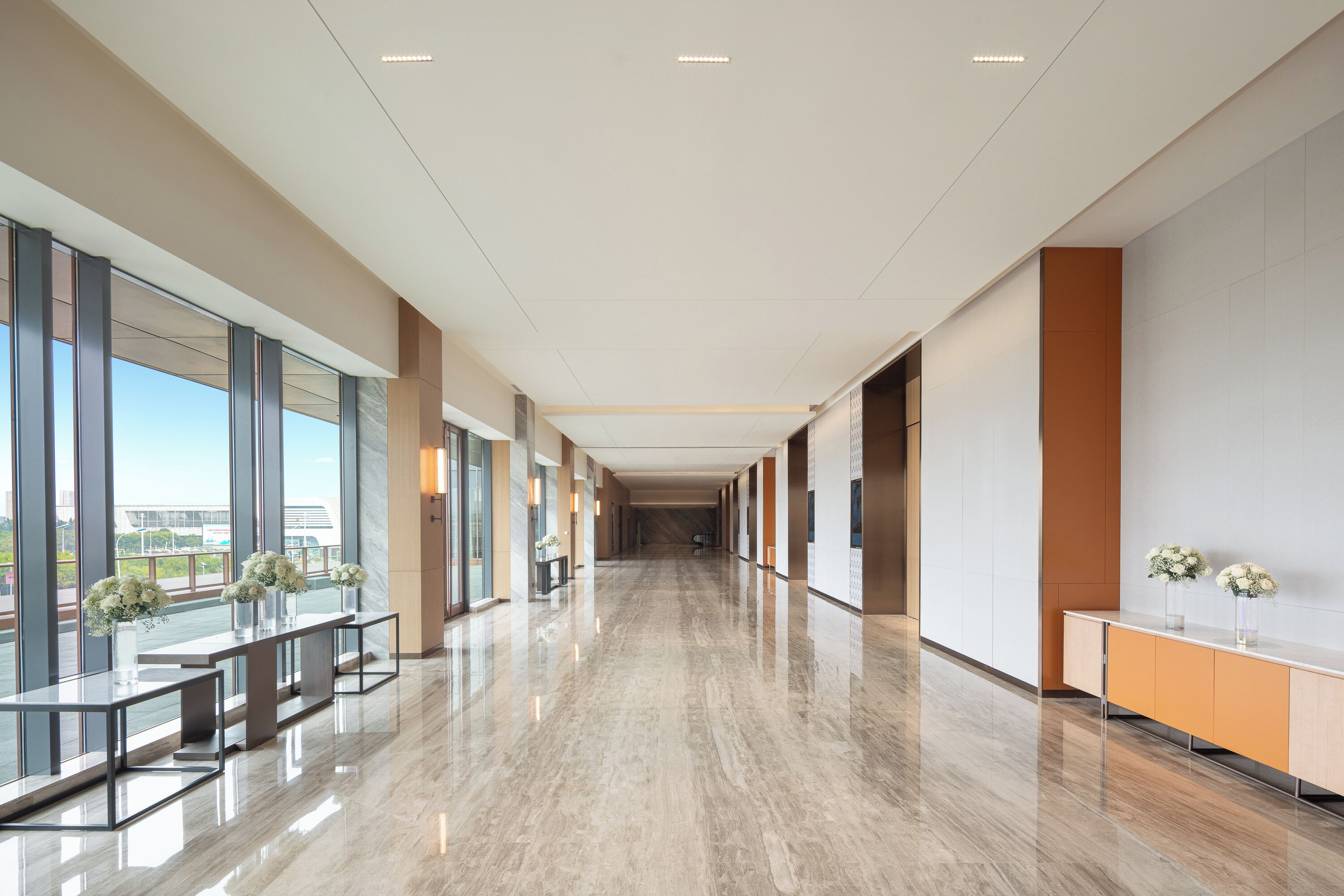 A modern hotel corridor with a polished marble floor, large windows, and a reception area with a counter and vases of flowers.