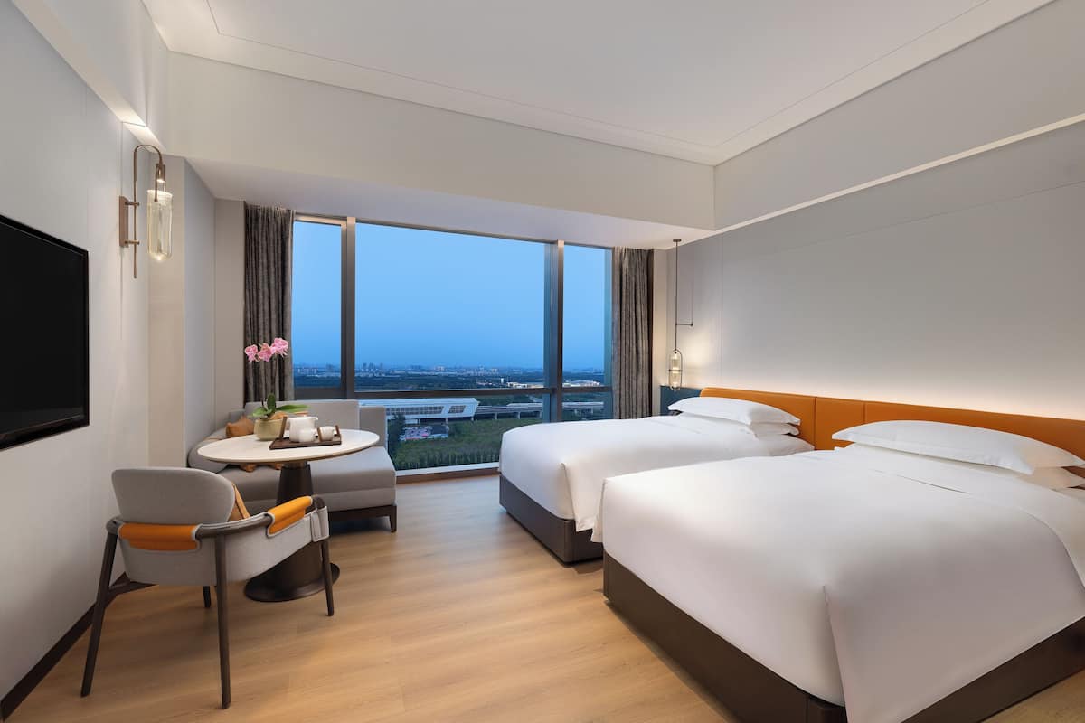 Een hotelkamer met twee bedden, een bureau, een stoel en een groot raam met uitzicht op de stad.