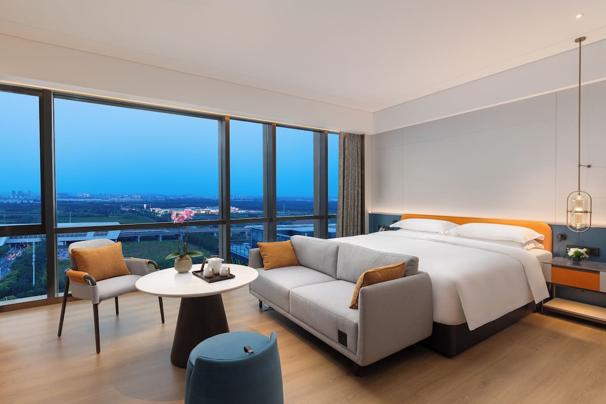 Een moderne hotelkamer met een groot bed, een bank, een kleine tafel en uitzicht op de skyline door grote ramen.
