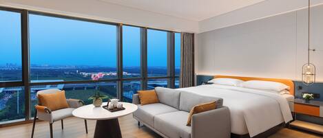 A modern hotel room with a large bed, a sofa, a small table, and a view of the cityscape through large windows.