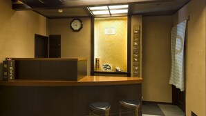 A reception area with a wooden counter, two black stools, a clock, and a wall with a wooden panel and a framed picture. - Kozuchi No Yado Tsurukame Daikichi (Nikko)