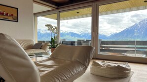 A modern living room with a leather sofa, a glass coffee table, and a potted orchid. A sliding glass door leads to a balcony with a mountain view. - die Villa", Eine Wohnung für 6 Personen (Sigriswil)