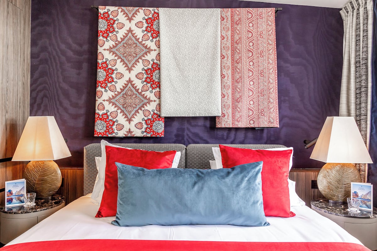 A hotel room with a bed, two nightstands, a lamp, a wall with patterned curtains, and a framed picture on the nightstand.