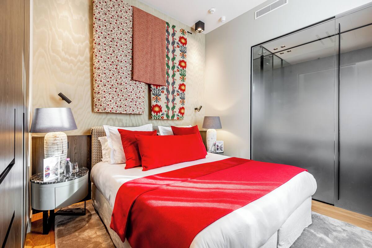 A modern hotel room with a large bed, a nightstand with a lamp, a wardrobe, and a wall with patterned wallpaper.