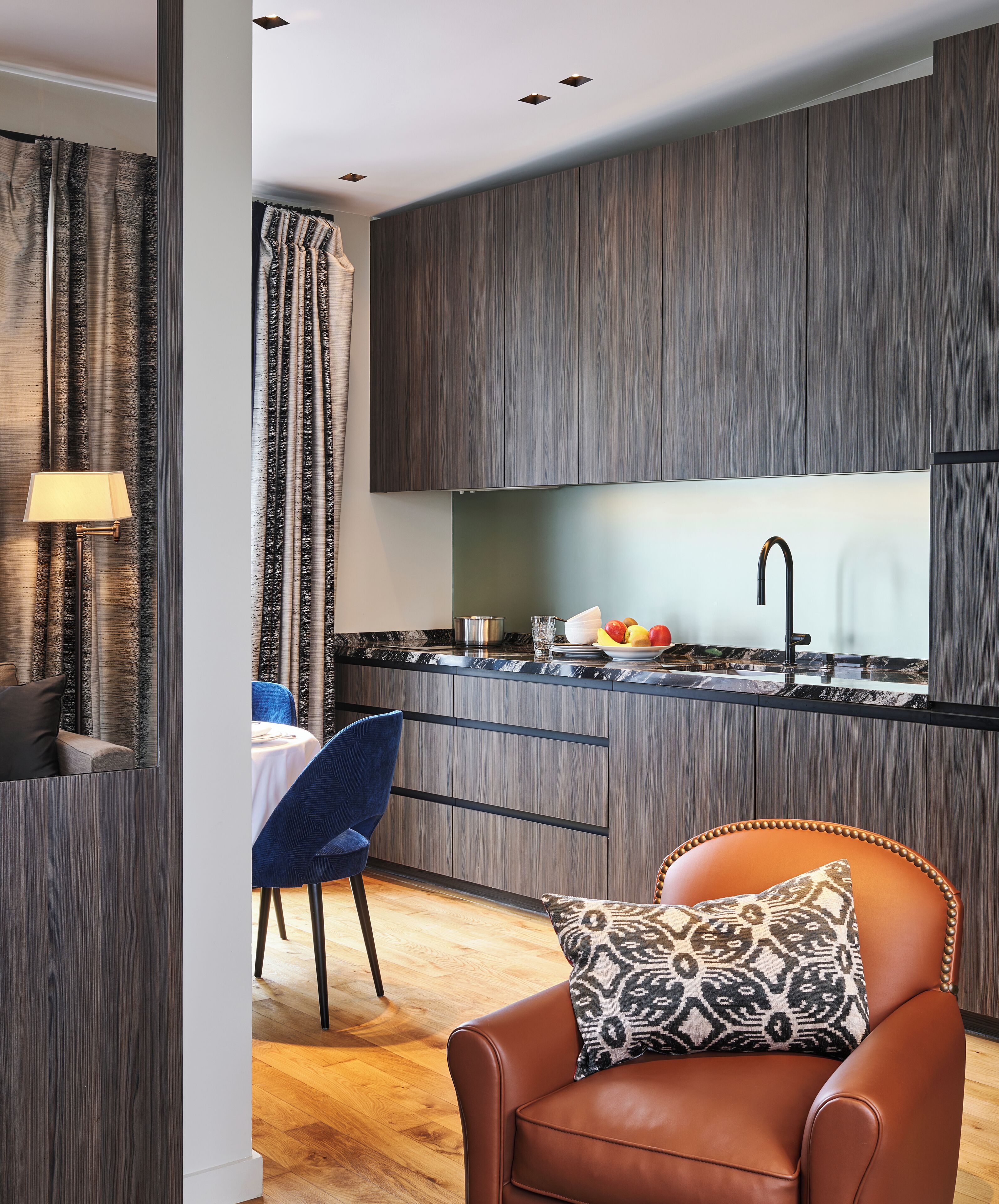 A modern kitchen with dark wood cabinets, a black faucet, and a marble countertop. There is a dining area with a blue chair and a brown leather sofa with a patterned pillow.