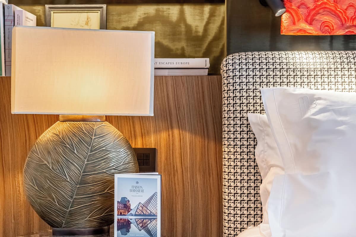 A neatly arranged hotel room with a bed, a bedside table with a lamp, a framed picture, and a bookshelf with books and decorative items.