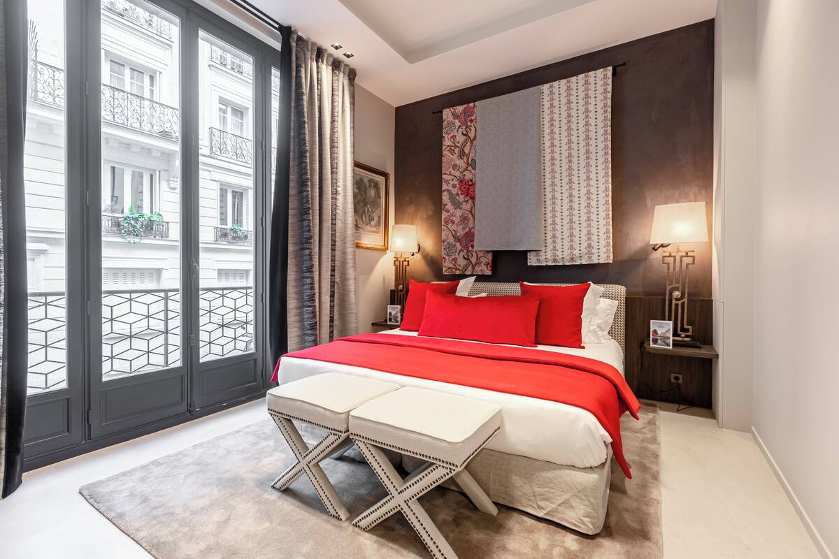 A modern bedroom with a large bed, a small table, two bedside lamps, and a view of the city through floor-to-ceiling windows.