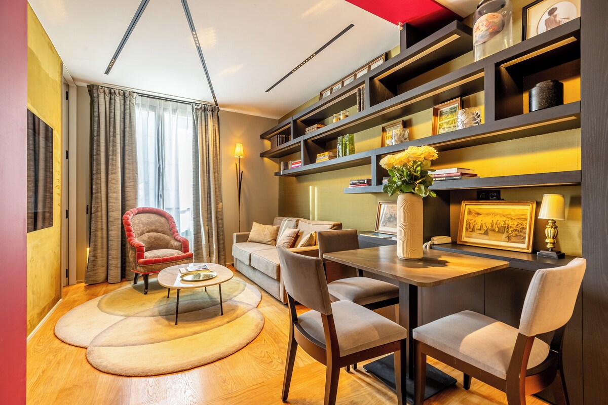 A modern living room with a sofa, armchairs, a coffee table, and a dining table with chairs. There are shelves with decorative items and a lamp.