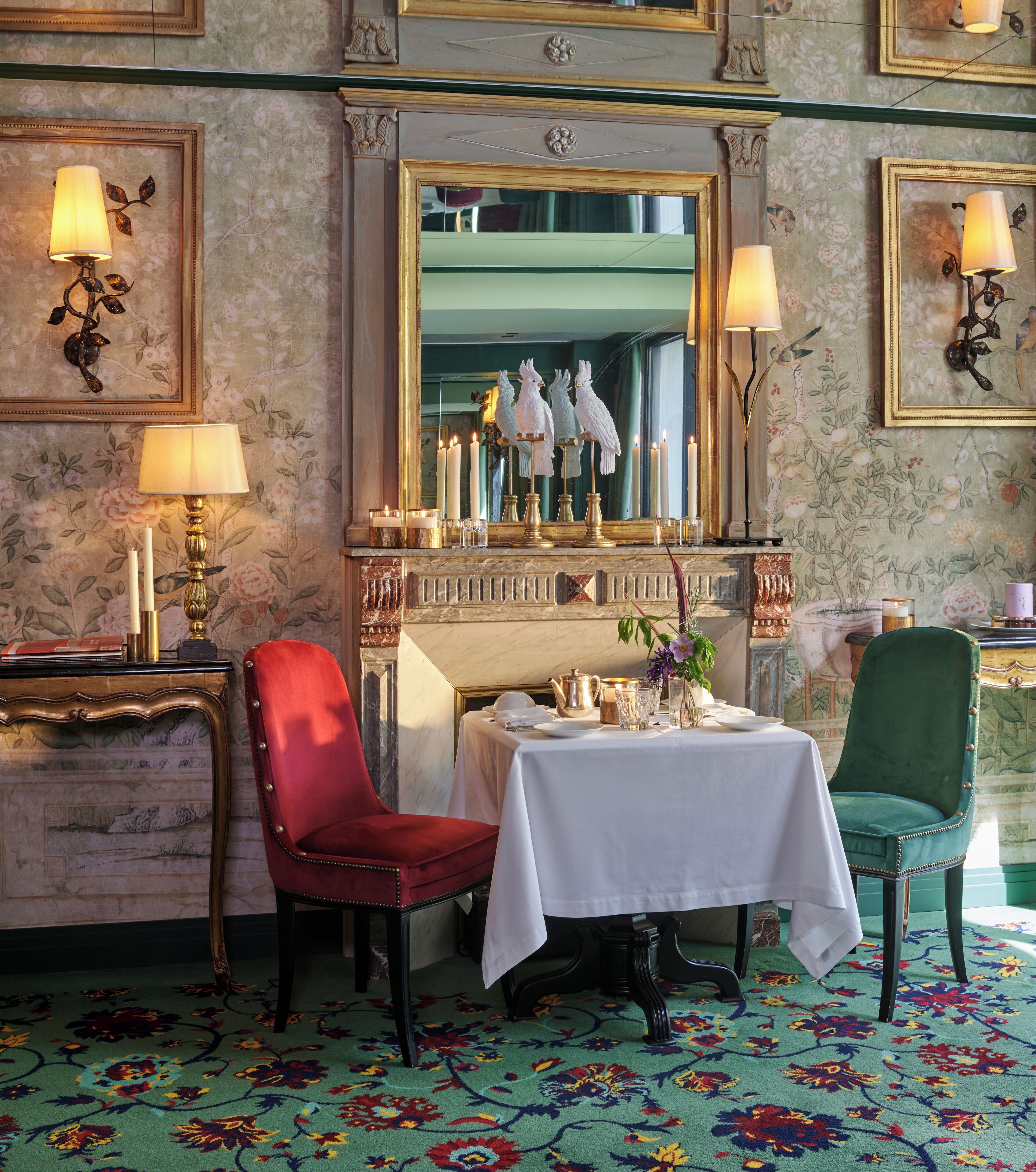 A dining area with a table set for two, a red chair, a green chair, and a mirror above the table.