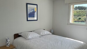 A bedroom with a bed, a bedside table, a framed picture on the wall, and a window with a view of greenery.