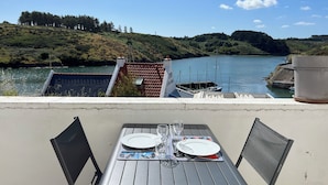 A table with plates and glasses set on a balcony overlooking a body of water with boats and a hilly landscape in the background.