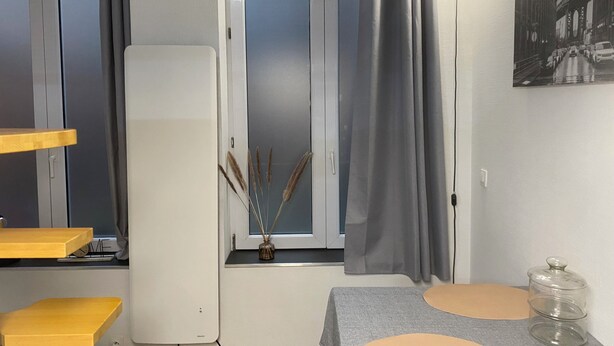 Une pièce avec une fenêtre, une table recouverte d’une nappe grise, un vase contenant des plantes séchées et un tableau encadré accroché au mur.