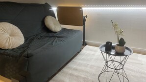 Un salon moderne avec un canapé gris foncé, une table ronde ornée d’un pot de plantes et une fenêtre illuminée par des lumières LED.