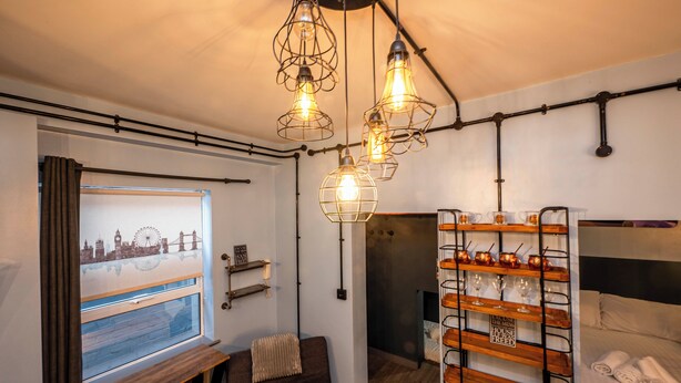 Une chambre d’hôtel compacte comprenant un lit, un coin salon avec un banc, une étagère fixée au mur avec des verres, et une fenêtre donnant sur le paysage urbain.