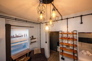 Una habitación de hotel compacta con una cama, una zona de estar con un banco, un estante empotrado con vasos y una ventana con vista a la ciudad.