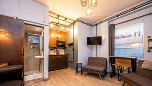 Un salon moderne comprenant un canapé, une petite table, un téléviseur fixé au mur et un coin cuisine avec un four à micro-ondes et des placards.