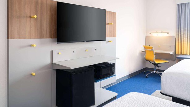 Habitación de hotel con televisión de pantalla plana montada en un panel de madera, un escritorio pequeño con refrigerador negro, una silla de oficina amarilla y una cama con edredón blanco.