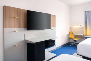 Ein Hotelzimmer mit Flachbildfernseher an einer Holzverkleidung, einem kleinen Schreibtisch mit einem schwarzen Kühlschrank, einem gelben Bürostuhl und einem Bett mit weißer Bettdecke.