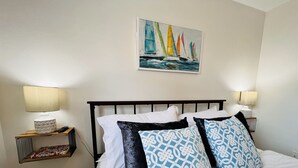 A neatly made bed with white and patterned pillows, a painting of sailboats on the wall, and two bedside lamps on wooden nightstands.