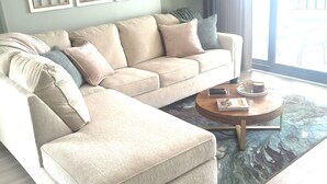 Un salon avec un canapé d’angle beige, une table basse ronde et une grande fenêtre avec des rideaux.