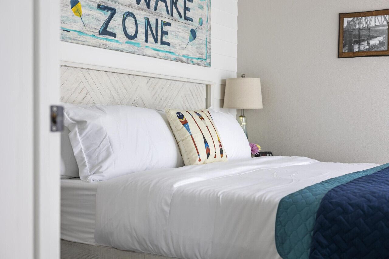 A neatly made bed with white linens and a teal quilted blanket, a decorative pillow with a colorful pattern, a wall sign that reads "ZONE," and a framed picture on the wall.