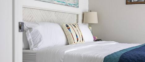 A neatly made bed with white linens and a teal quilted blanket, a decorative pillow with a colorful pattern, a wall sign that reads "ZONE," and a framed picture on the wall.