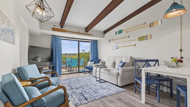 A modern living room with a sofa, chairs, a dining table, and a TV. There are wooden beams on the ceiling, a large window with curtains, and a view of greenery outside. - Pool, Pickleball, Hot Tub, Lakeview- Lakecation (Kimberling City)