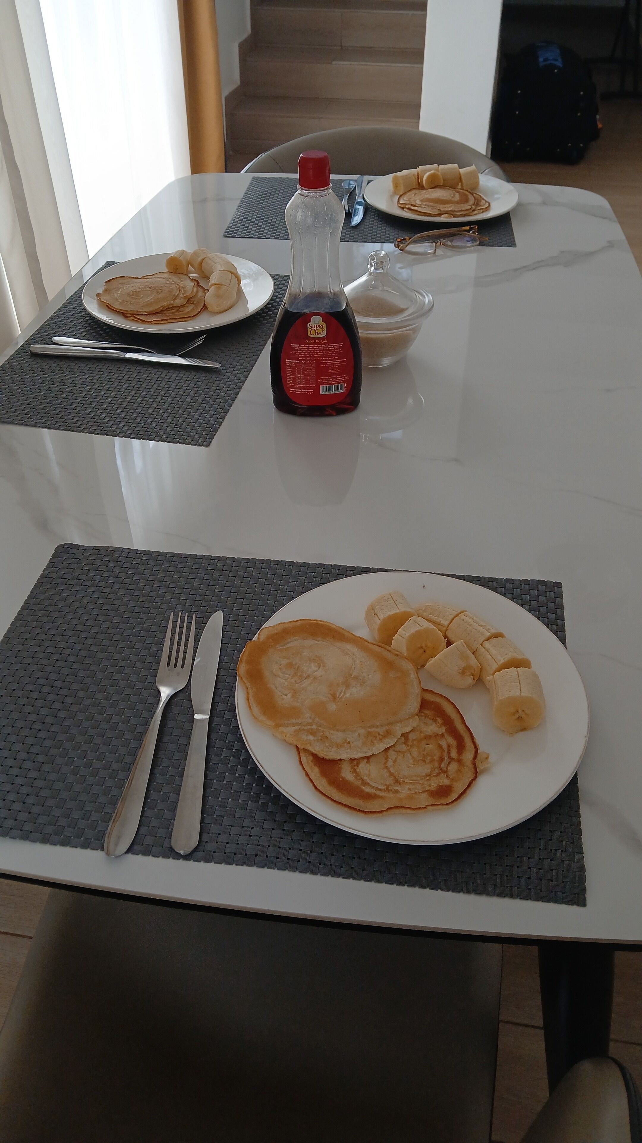 Una mesa de comedor con platos de tortitas y plátanos, una botella de sirope y cubiertos.