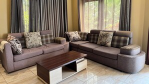A living room with a sectional sofa, a wooden coffee table, and a large window with curtains. - Dafrican BnB  (Kigali)