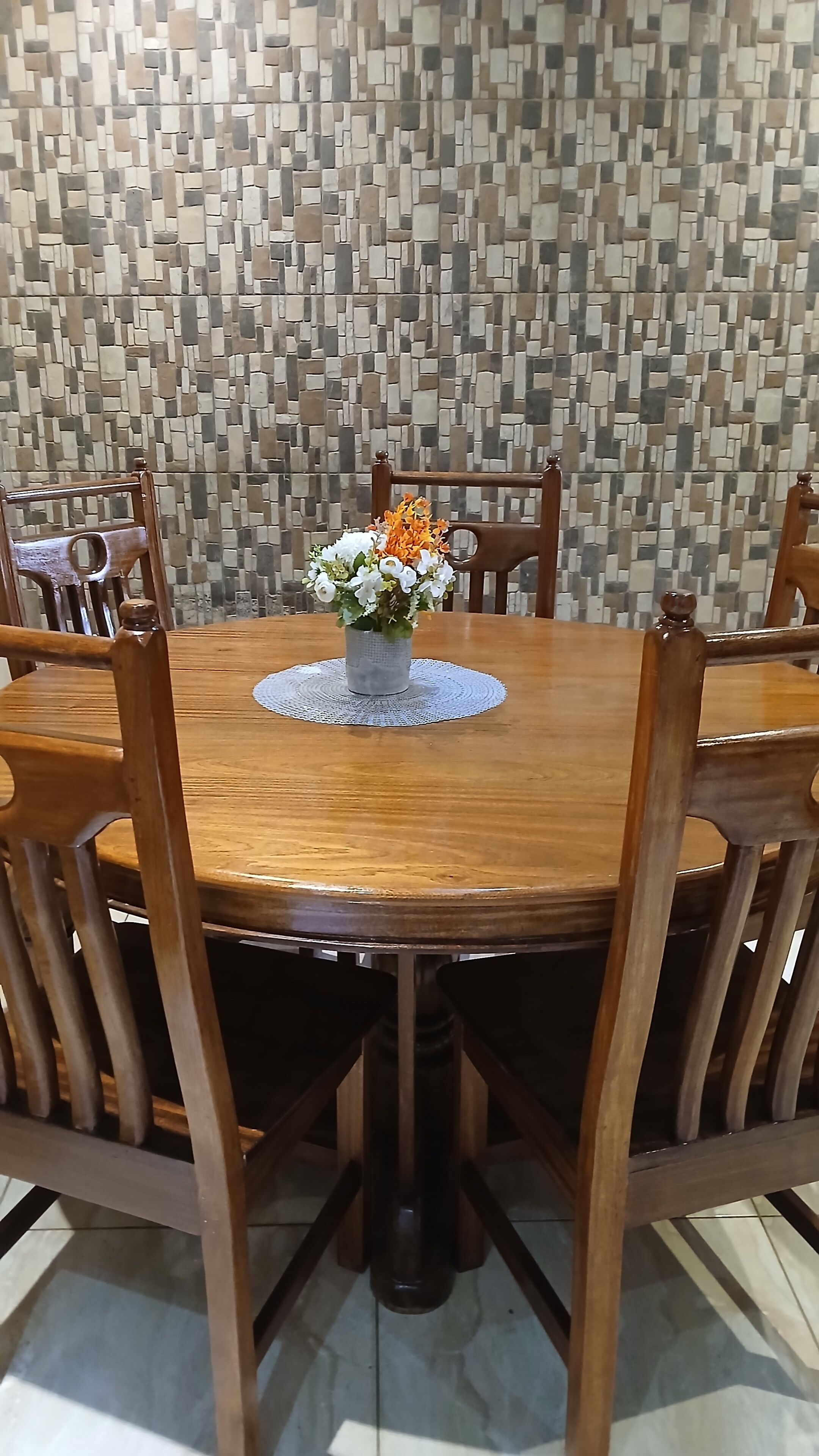 Une table de salle à manger ronde en bois, surmontée d’un set de table en fibres tressées, est entourée de chaises en bois. Un arrangement floral est placé au centre de la table.