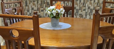 A round wooden dining table with a lace doily, surrounded by wooden chairs with cut-out backrests. In the center, there is a vase with flowers. The background features a mosaic wall.