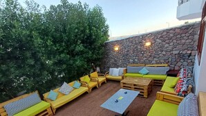 Un espacio al aire libre con mobiliario de madera y cojines, una pared de piedra y un árbol de fondo.