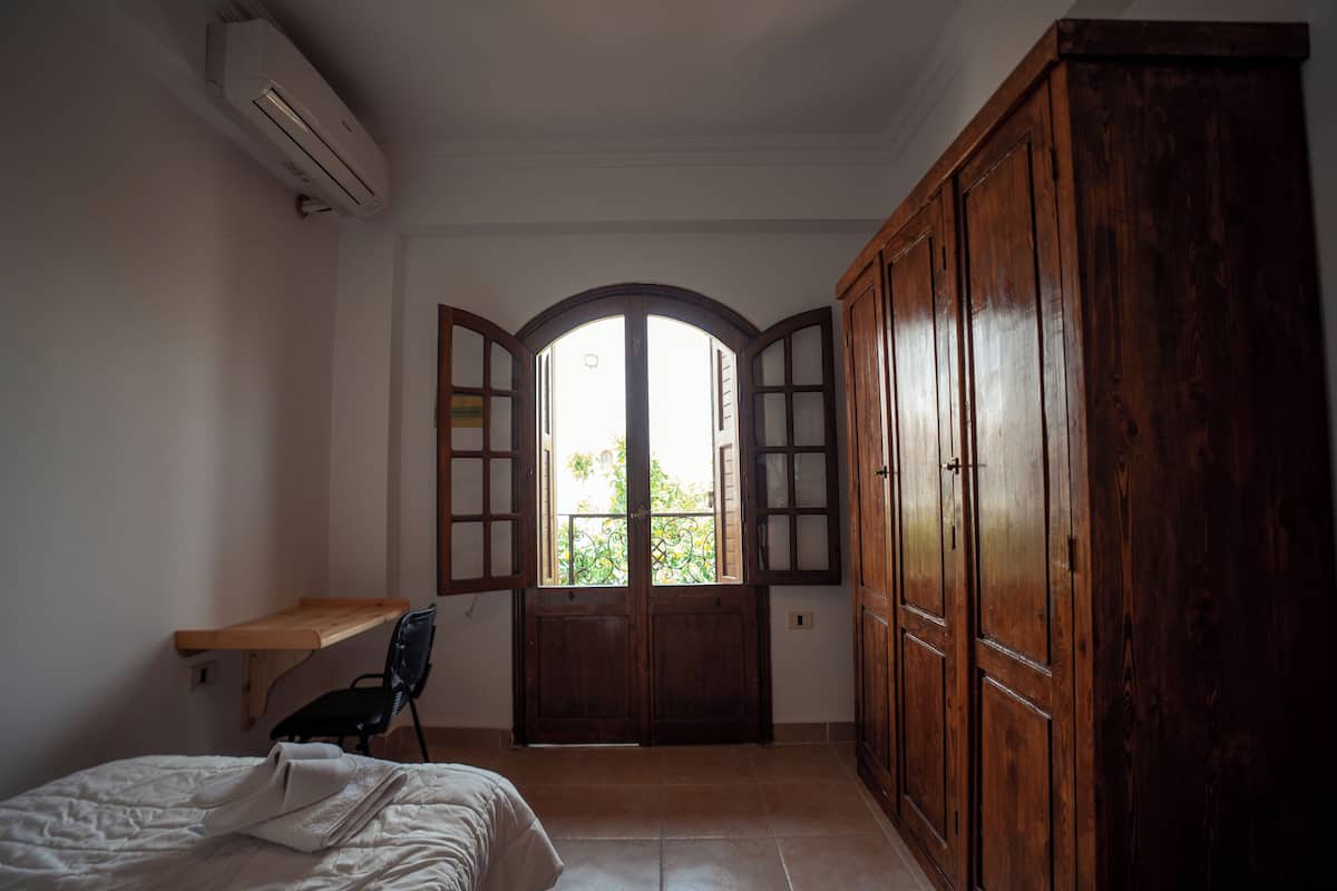 Un dormitorio con cama, un escritorio de madera, una silla, un armario y una ventana con contraventanas abiertas.